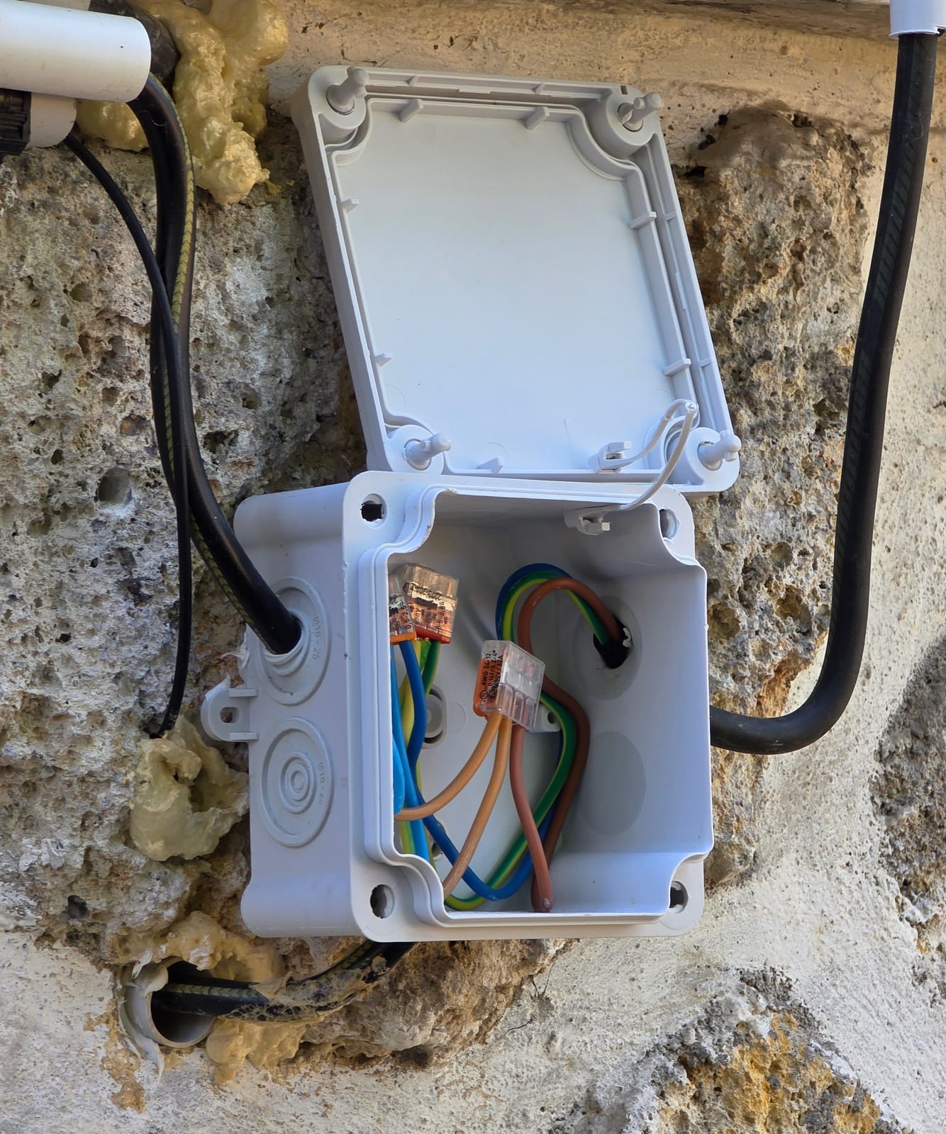 Scatola di derivazione esterna aperta su un muro in pietra con cavi, connettori e schiuma espansiva