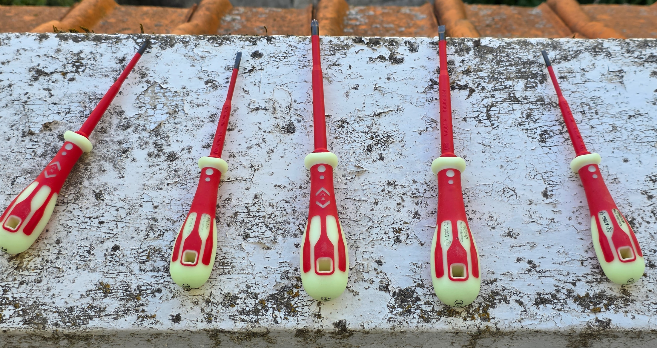 Cinque cacciaviti VDE C.K Tools con manici rossi e fosforescenti allineati su un muretto bianco all’aperto