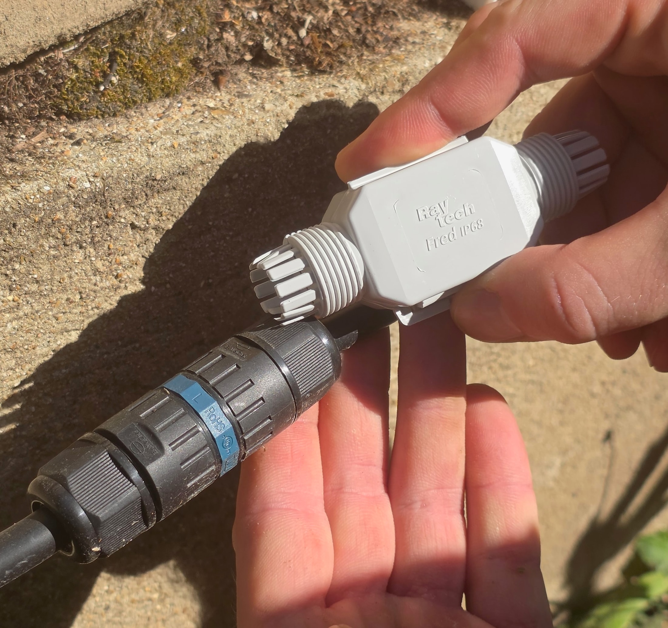 Grauer wasserdichter Steckverbinder Raytech Fred IP68 in der Hand gehalten, gegenüber einem daneben liegenden schwarzen Steckverbinder