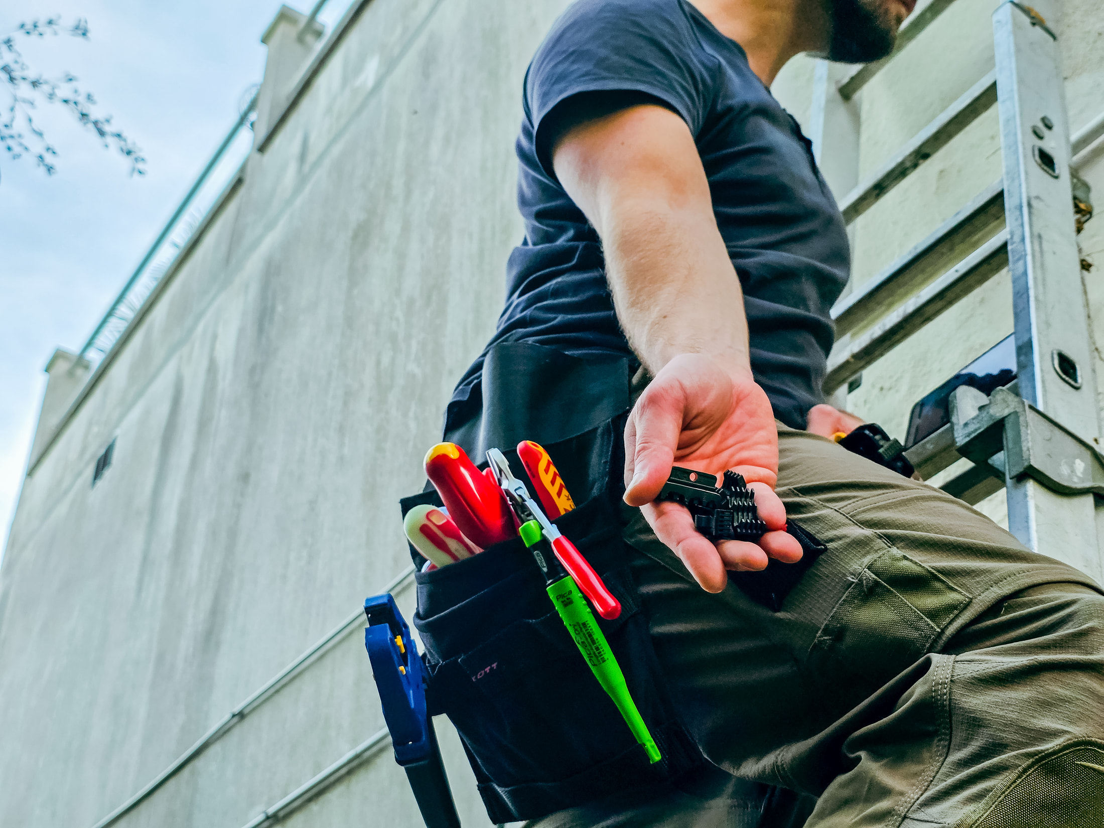 A black fixing clip held in the hand, a belt loaded with red-handled tools and a green tester, and a pair of work trousers.