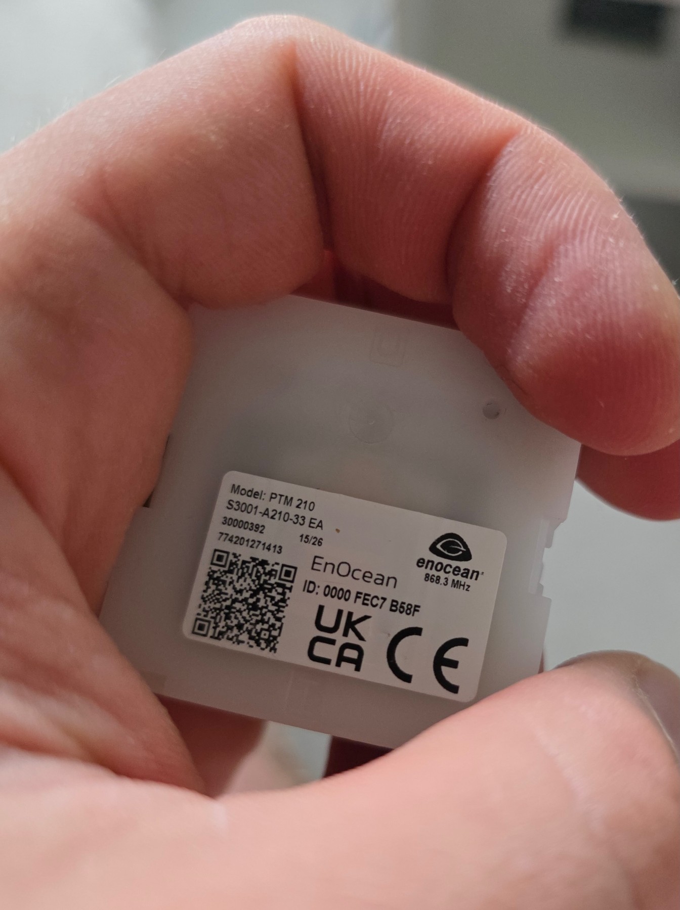 Dorso di un modulo radio EnOcean PTM 210 tenuto in mano, con un’etichetta che riporta il modello, l’identificativo radio e la frequenza 868,3 MHz.