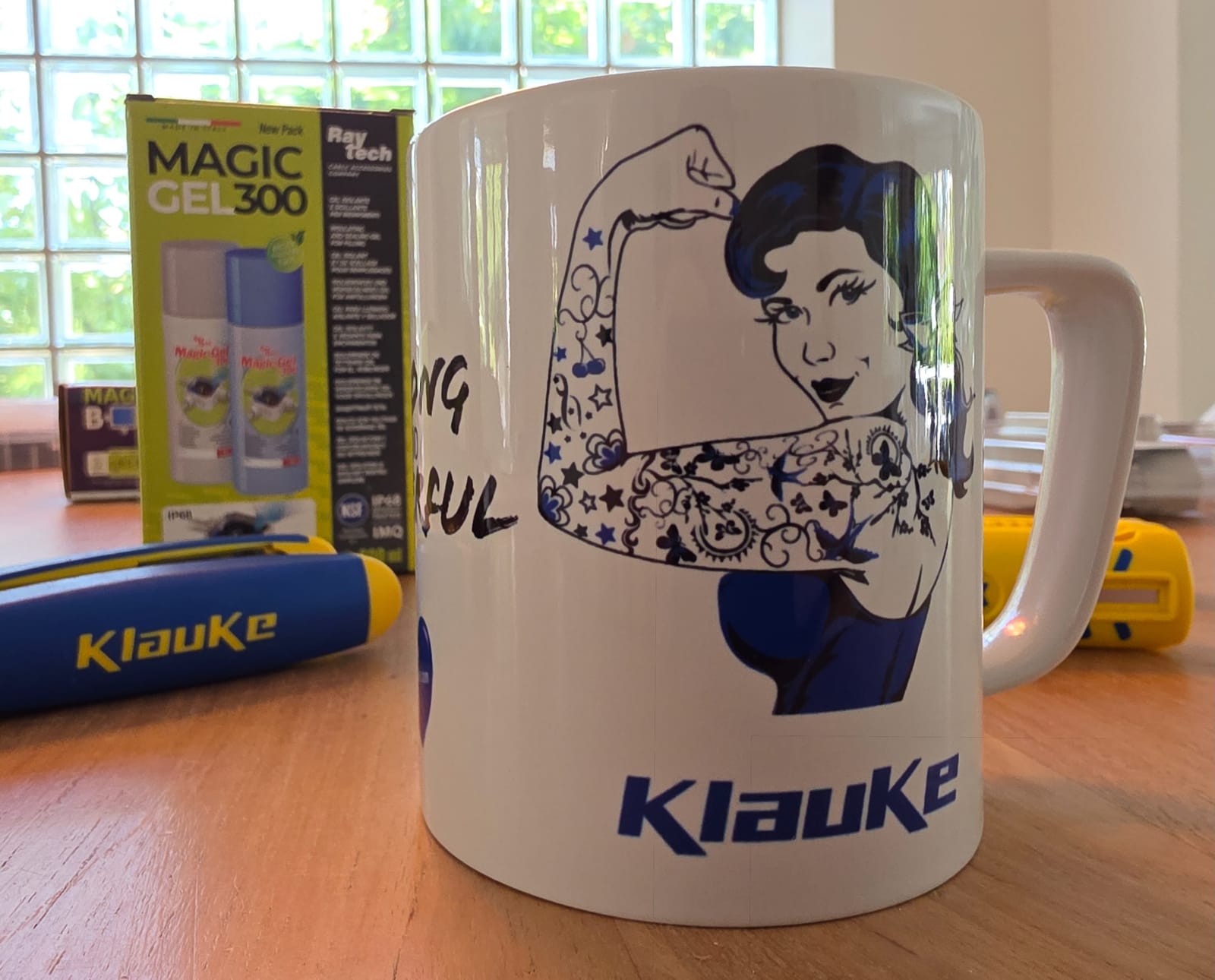 Taza blanca Klauke con un dibujo de una mujer con el brazo levantado, una caja de gel Raytech al fondo