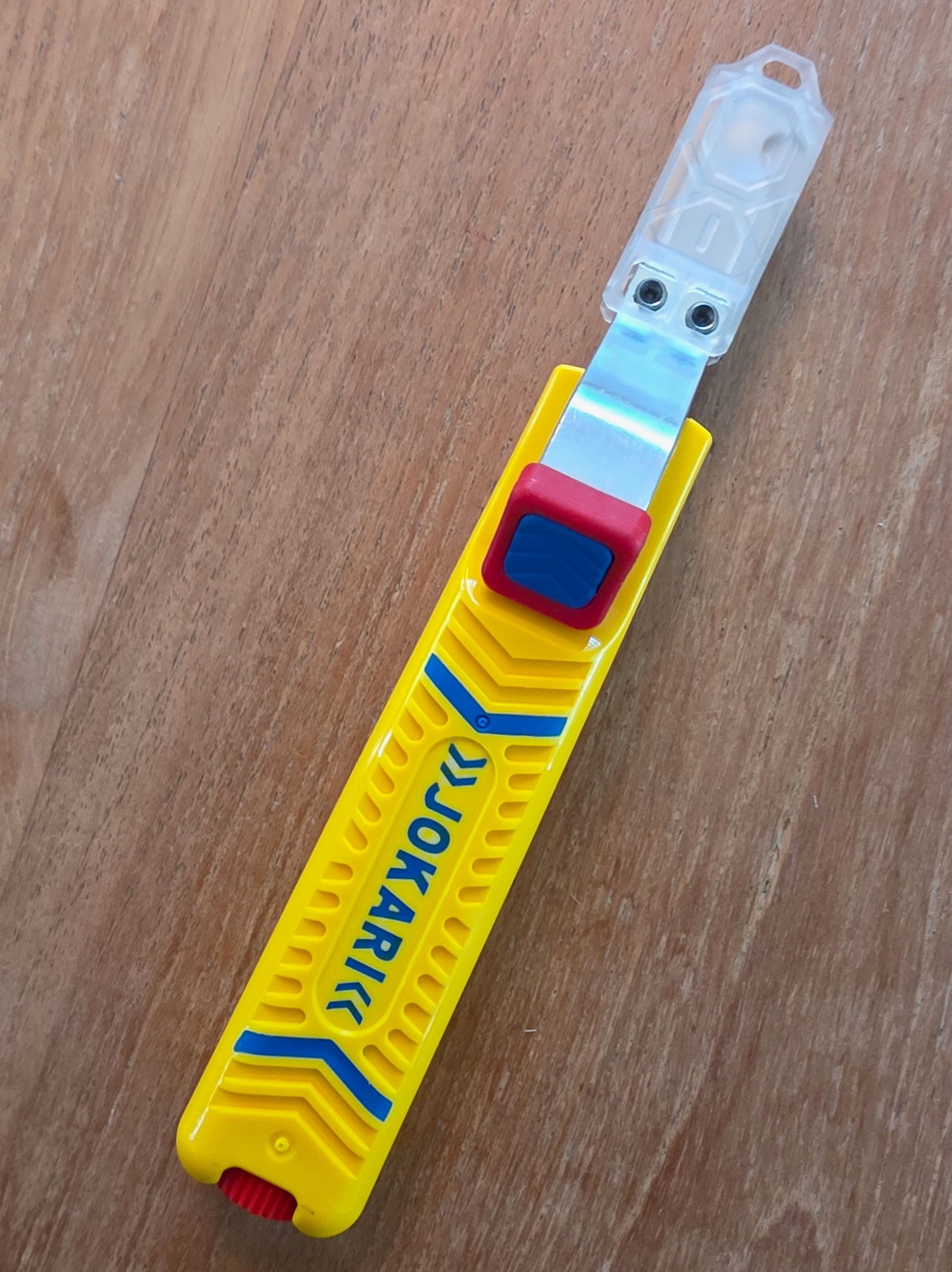 Coltello spellacavi Jokari No. 28H fuori dalla confezione, appoggiato su un tavolo di legno, lama a uncino aperta con il suo cappuccio di protezione