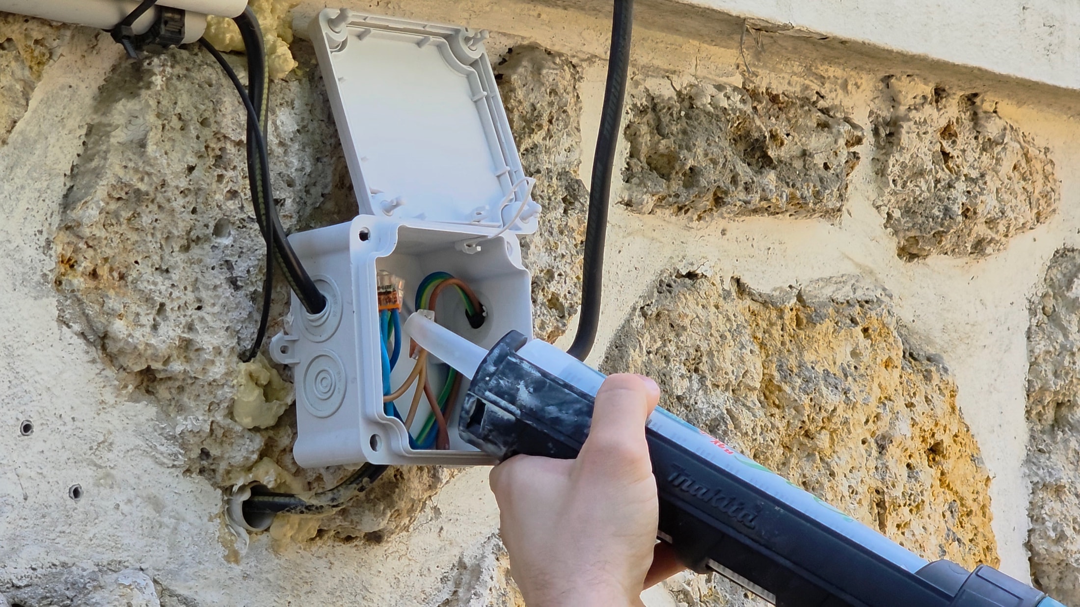 Pistola per silicone Makita che applica il gel isolante in una scatola di derivazione su un muro in pietra