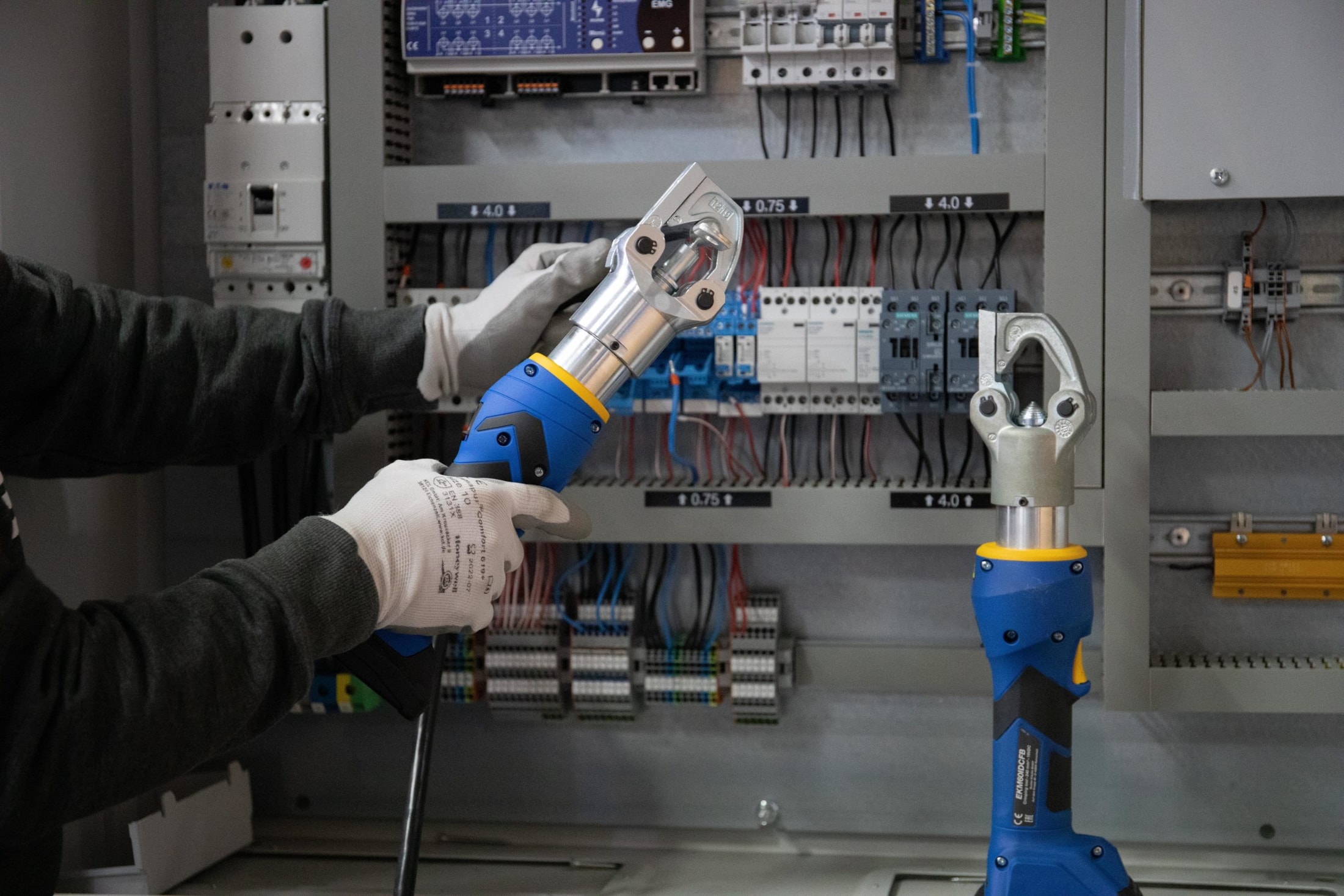 Un elettricista con i guanti utilizza una crimpatrice elettrica blu e nera davanti a un quadro elettrico aperto, con una seconda crimpatrice identica posata accanto.
